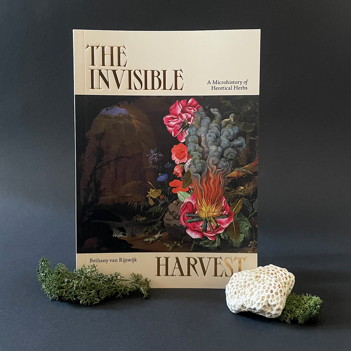 A book titled 'THE INVISIBLE HARVEST' with a floral and fungi artwork cover, placed on a dark surface alongside some natural elements.