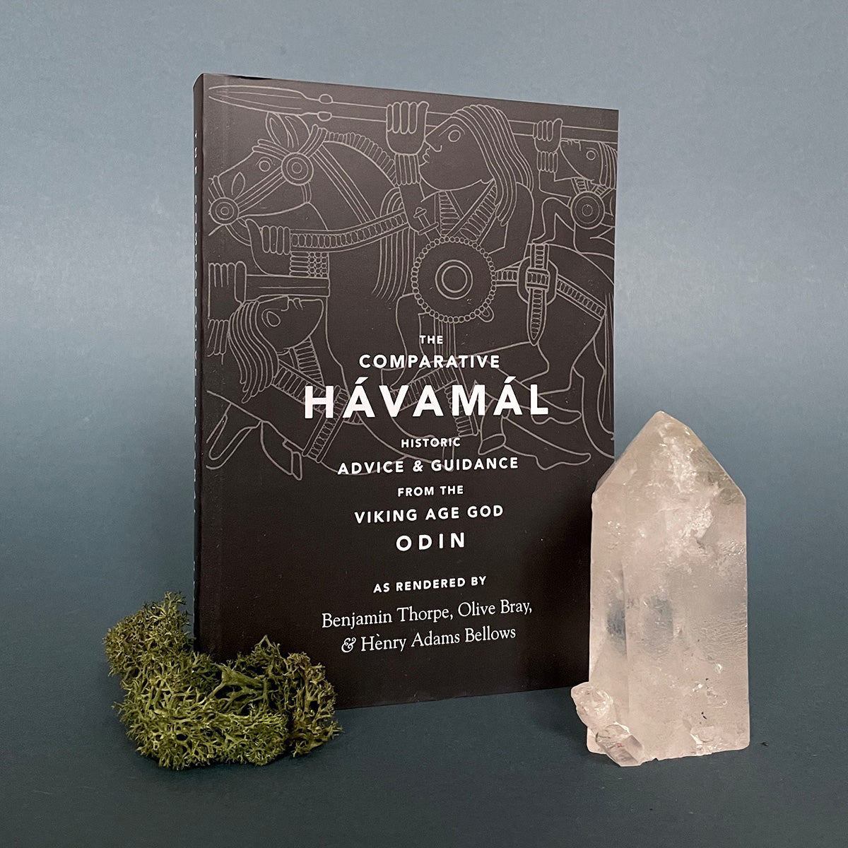 A book titled 'The Comparative Hávamál" featuring a black cover with white text and a symbol design. The book is presented next to a crystal and a pine cone on a dark background.