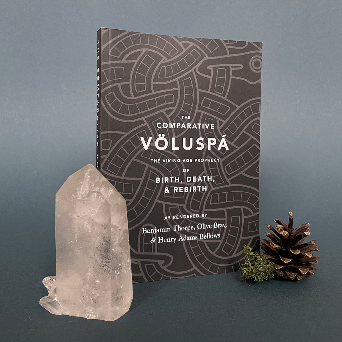 A book titled 'The Comparative Völuspá featuring a black cover with white text and a symbol design. The book is presented next to a crystal and a pine cone on a dark background.