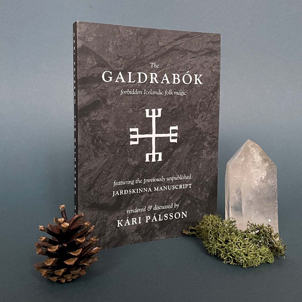 A book titled 'The Galdrabók: forbidden Icelandic folk magic' by Kári Pálsson, featuring a black cover with white text and a symbol design. The book is presented next to a crystal and a pine cone on a dark background.