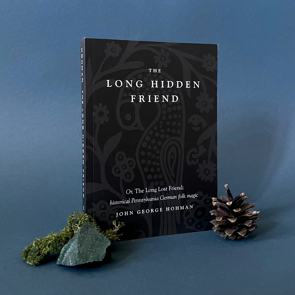 A book titled 'The Long Hidden Friend' by John George Hohman, presented on a dark background with a decorative pattern on the cover, accompanied by a pine cone and a piece of moss.
