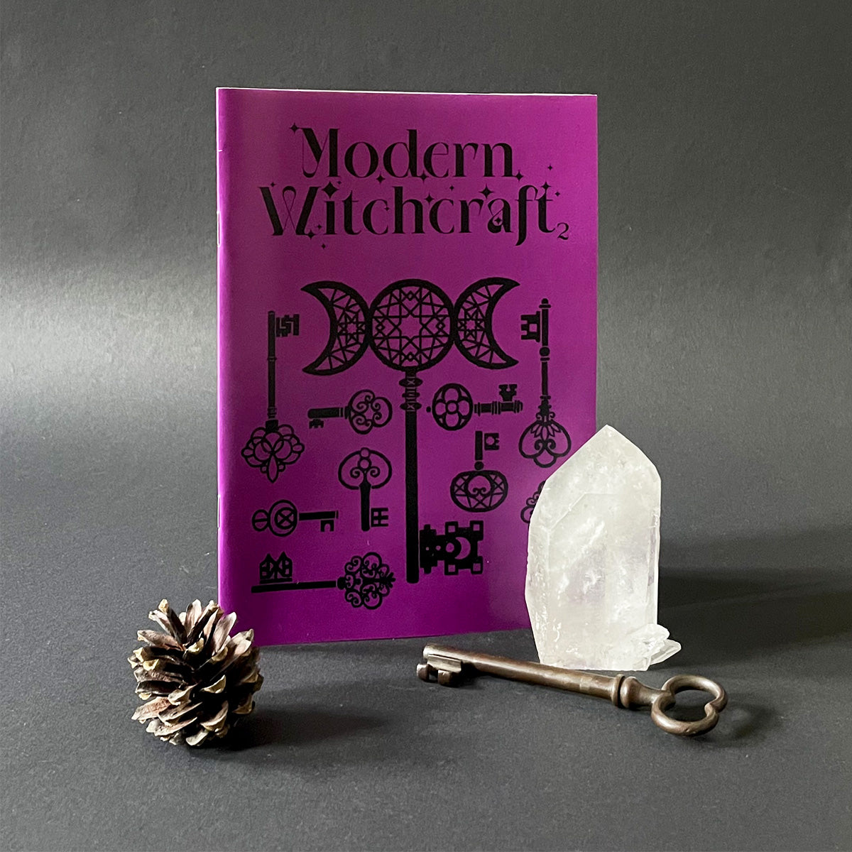 A purple magazine titled 'Modern Witchcraft' with a graphic design, accompanied by a white crystal and an metallic key on a gray background.