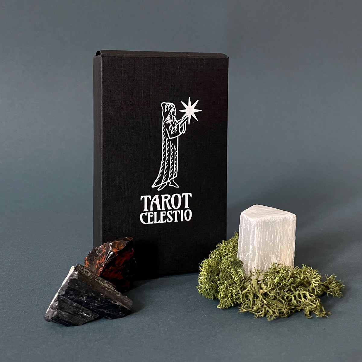 A black Tarot Celestio tuck box with white foil embossing of a figure holding a star, a piece of black stone, and a bundle of moss on a gray background.