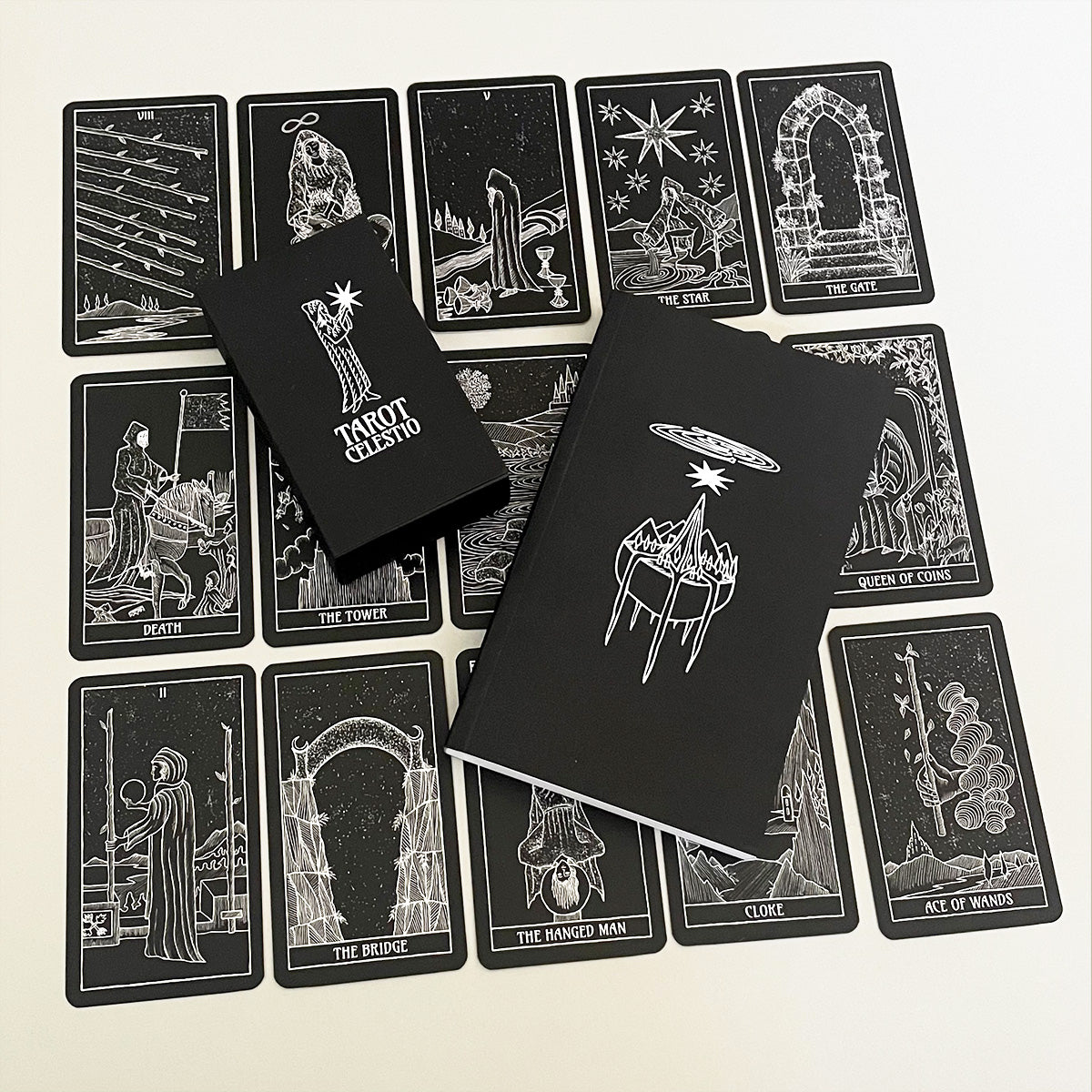 A collection of black and white tarot cards with intricate pen and ink illustrations laid out on a surface, along with a black tuck box and a black book with white text.
