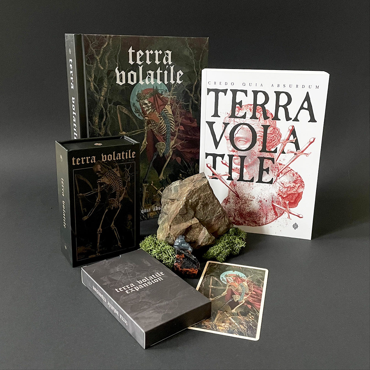 Collection of 'Terra Volatile' components including books, box, and cards on a dark background