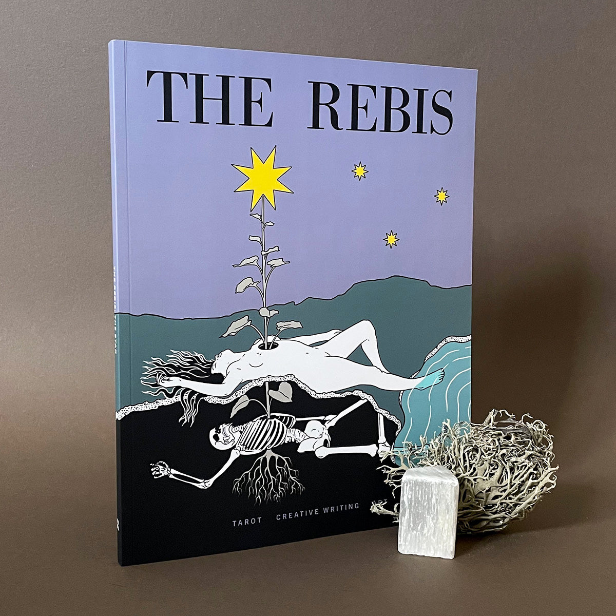 A magazine titled 'THE REBIS' with a star-themed cover design featuring illustrations of stars, mountains, and a human figure. The book is accompanied by a small, decorative item that resembles a woven basket or sculpture.