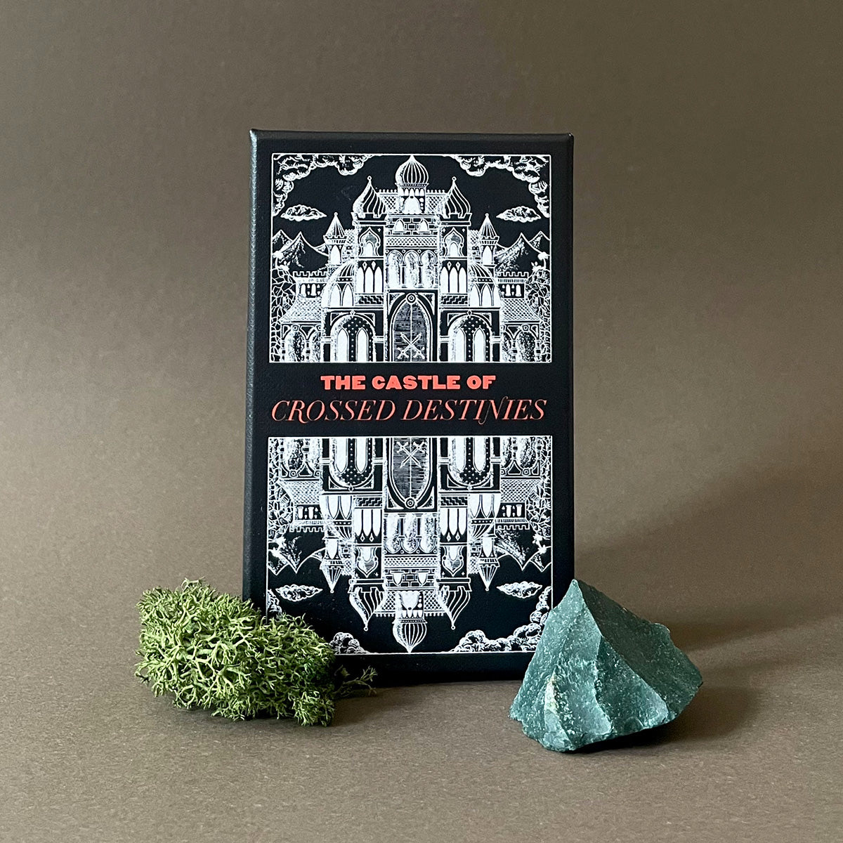 A tarot deck titled 'The Castle of Crossed Destinies' displayed with its artwork featuring a castle on the cover, accompanied by a green crystal and a sprig of moss.