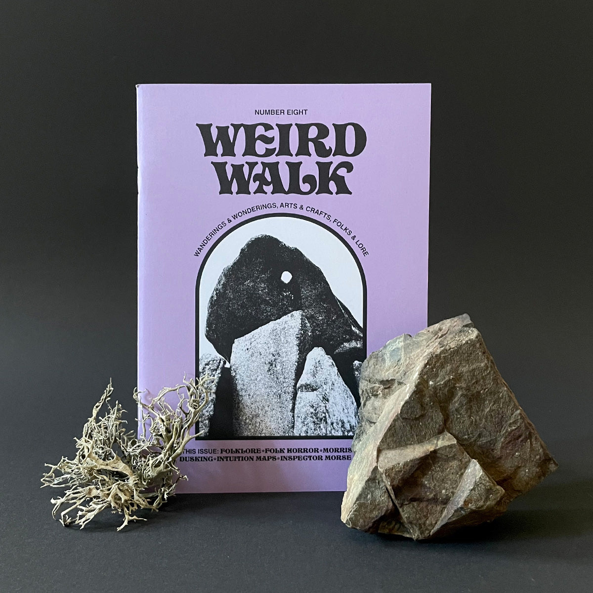 A cover image of 'Weird Walk Issue 8' magazine with a graphic of a standing stone, accompanied by a natural rock and dried plant decoration.
